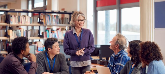 Teacher With Group Of Mature Adult Students In Class Sit Around Table And Work In College Library